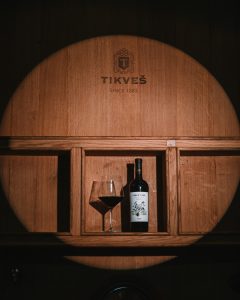 čaša crvenog vina Luda Mara Red Cuvée Tikveć, na drvenoj podlozi i drvenim buretom u pozadini, za svetski dan vranca