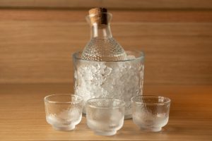 sake u boci koja se hladi u posudi s ledom, ispred su tri čaše, sve od debljeg matiranog stakla sa drvenom pozadinom i barskim drvenim stolom u naturalnim tonovima